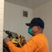 A worker wearing an orange long-sleeve shirt, dark blue baseball cap, and teal respiratory mask is using a DeWalt rotary hammer drill on a white-painted concrete block wall. The setting appears to be an interior room undergoing renovation or construction work. Dust and debris are visible at the base of the wall where drilling is occurring.