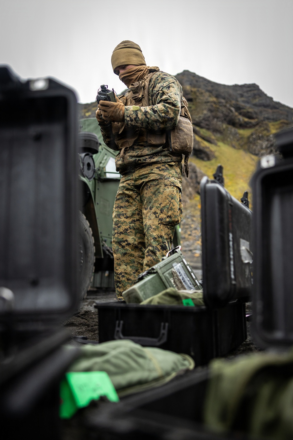A military service member in MARPAT (Marine pattern) digital camouflage uniform stands outdoors in a cold, overcast environment, examining a handheld device. The setting appears to be a rocky, mountainous coastal or northern terrain with military vehicles and equipment cases visible nearby. The scene suggests a field operation or exercise in a cold-weather environment.