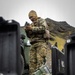 A military service member in MARPAT (Marine pattern) digital camouflage uniform stands outdoors in a cold, overcast environment, examining a handheld device. The setting appears to be a rocky, mountainous coastal or northern terrain with military vehicles and equipment cases visible nearby. The scene suggests a field operation or exercise in a cold-weather environment.