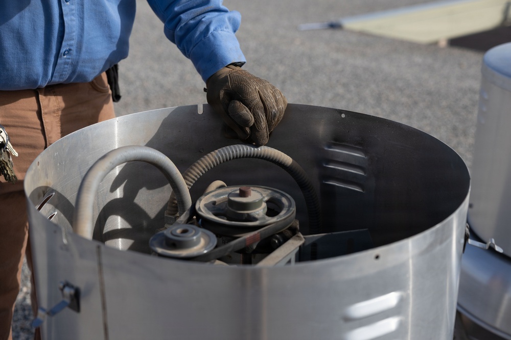 A person wearing a blue shirt, brown pants, and dark work gloves is handling the lid or cover of a large cylindrical metal container. Inside the container, mechanical components are visible including pulleys, belts, and a flexible hose. The setting appears to be an outdoor paved area, likely a parking lot or work site.