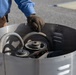 A person wearing a blue shirt, brown pants, and dark work gloves is handling the lid or cover of a large cylindrical metal container. Inside the container, mechanical components are visible including pulleys, belts, and a flexible hose. The setting appears to be an outdoor paved area, likely a parking lot or work site.