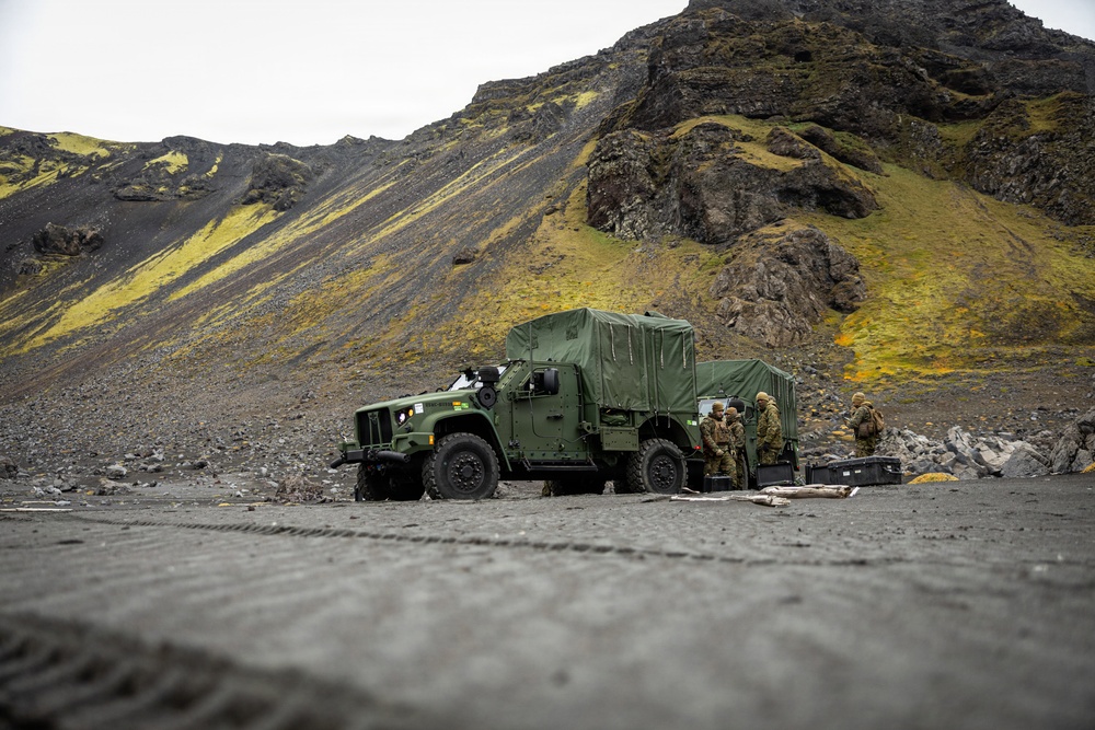 Military personnel are gathered beside a green tactical vehicle, likely an Oshkosh JLTV or similar MRAP-class truck, in a rugged volcanic terrain. The landscape features dark basaltic gravel and moss-covered rocky hillsides consistent with Icelandic or similar North Atlantic geography. Several soldiers in combat uniforms appear to be conducting equipment checks or a briefing near the vehicle.