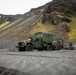 Military personnel are gathered beside a green tactical vehicle, likely an Oshkosh JLTV or similar MRAP-class truck, in a rugged volcanic terrain. The landscape features dark basaltic gravel and moss-covered rocky hillsides consistent with Icelandic or similar North Atlantic geography. Several soldiers in combat uniforms appear to be conducting equipment checks or a briefing near the vehicle.