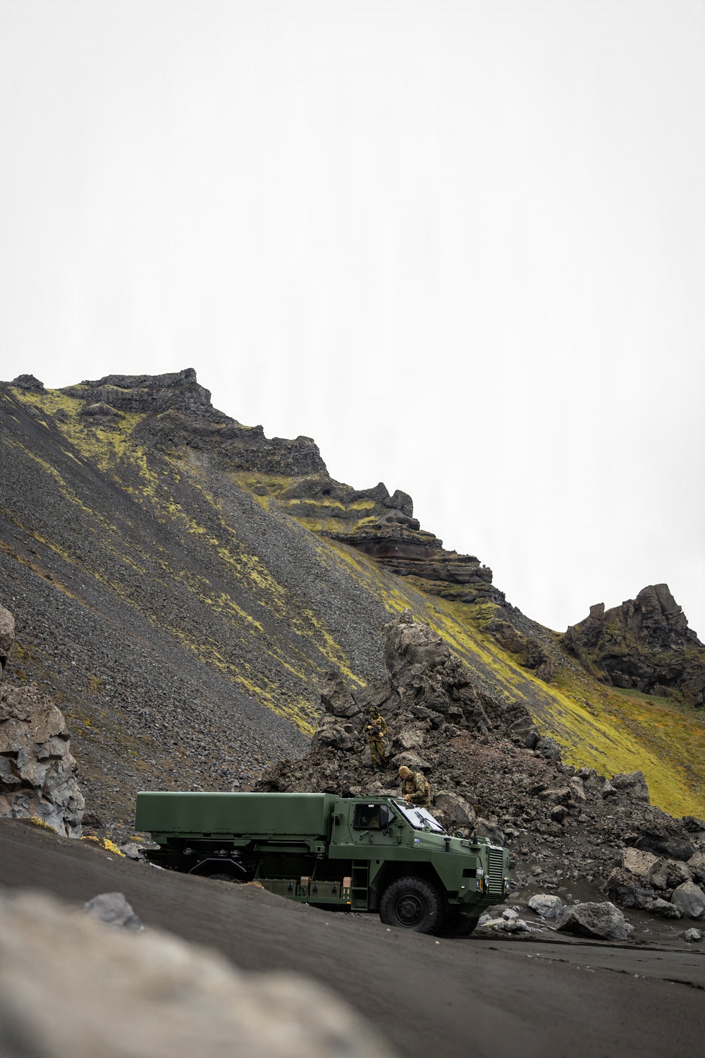 A military green cargo truck is positioned on a dark volcanic terrain road at the base of a steep, rocky hillside with patches of yellow-green moss or lichen. Two personnel in camouflage uniforms are visible near the front cab area of the vehicle. The landscape strongly suggests an Icelandic or similarly volcanic North Atlantic environment.