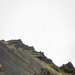 A military green cargo truck is positioned on a dark volcanic terrain road at the base of a steep, rocky hillside with patches of yellow-green moss or lichen. Two personnel in camouflage uniforms are visible near the front cab area of the vehicle. The landscape strongly suggests an Icelandic or similarly volcanic North Atlantic environment.