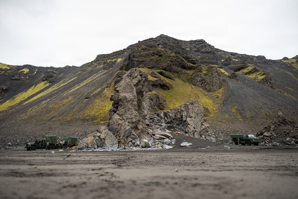 A dramatic volcanic landscape featuring dark basaltic mountains with patches of yellow-green moss or lichen, likely in Iceland or a similar subarctic environment. Several military vehicles in olive-green camouflage are parked near a prominent rock formation at the base of the mountain. A small number of personnel in military uniforms are visible near the vehicles and rock outcrop.