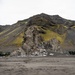 A dramatic volcanic landscape featuring dark basaltic mountains with patches of yellow-green moss or lichen, likely in Iceland or a similar subarctic environment. Several military vehicles in olive-green camouflage are parked near a prominent rock formation at the base of the mountain. A small number of personnel in military uniforms are visible near the vehicles and rock outcrop.