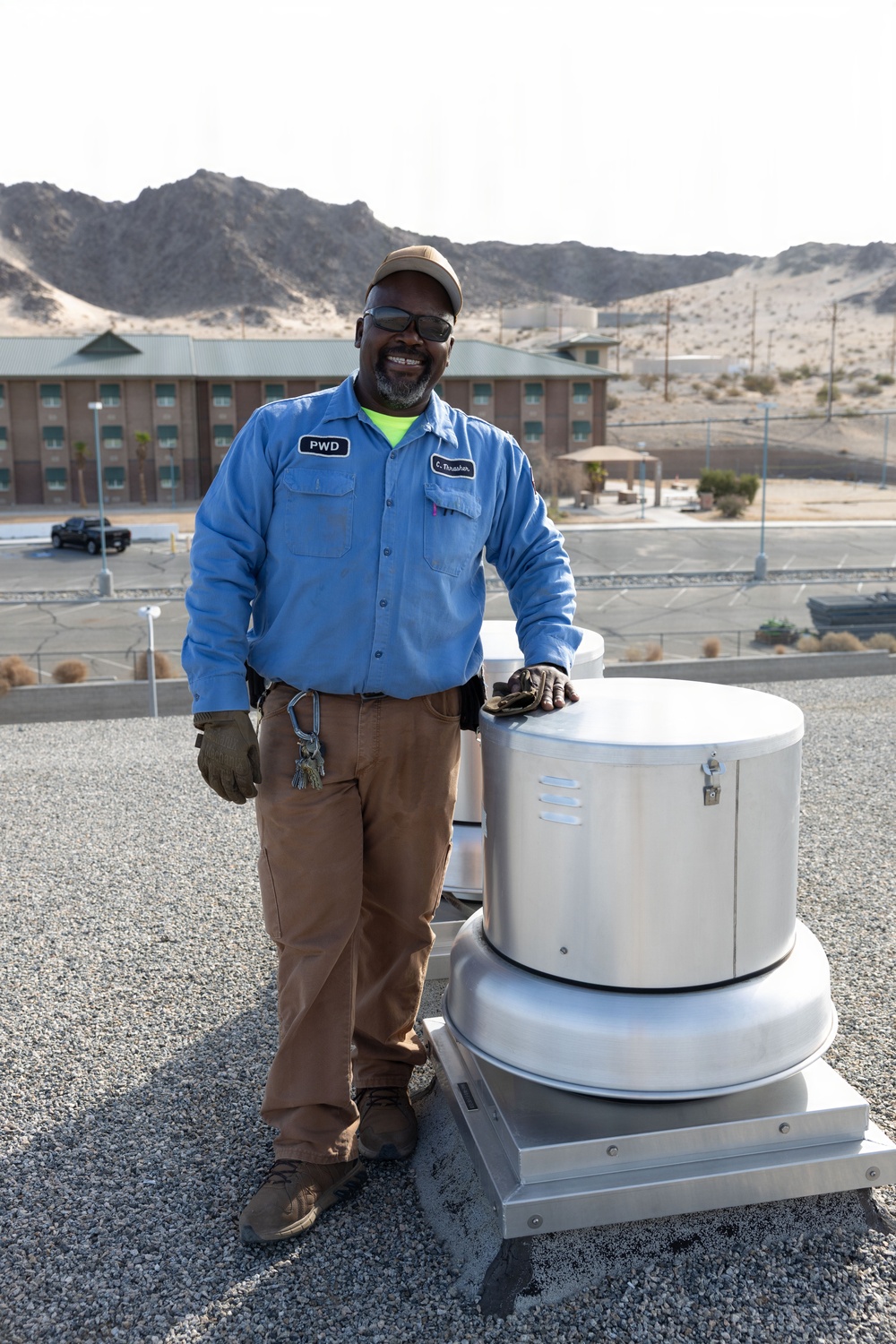 A maintenance worker wearing a blue uniform shirt with 'PWD' and a name tag reading 'C. Thrasher' stands on a flat rooftop next to a rooftop exhaust fan or ventilation unit. He is smiling, wearing sunglasses and a tan cap, with work gloves and keys visible. The background shows a desert landscape with rocky mountains, a multi-story building, and a parking area.