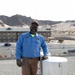 A maintenance worker wearing a blue uniform shirt with 'PWD' and a name tag reading 'C. Thrasher' stands on a flat rooftop next to a rooftop exhaust fan or ventilation unit. He is smiling, wearing sunglasses and a tan cap, with work gloves and keys visible. The background shows a desert landscape with rocky mountains, a multi-story building, and a parking area.