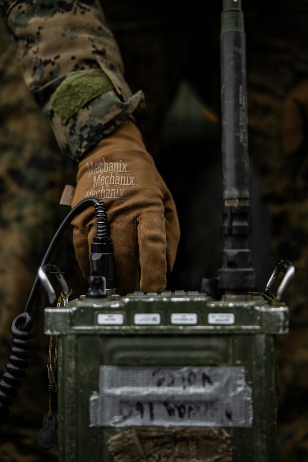 A military service member wearing MARPAT digital camouflage and Mechanix brand tactical gloves is operating a military field radio set, identifiable by its olive drab casing, antenna, and control panel. The equipment appears to be a ruggedized military communications device with visible labels and a coiled handset cable. The close-up composition emphasizes the hands-on interaction with the radio equipment.