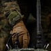 A military service member wearing MARPAT digital camouflage and Mechanix brand tactical gloves is operating a military field radio set, identifiable by its olive drab casing, antenna, and control panel. The equipment appears to be a ruggedized military communications device with visible labels and a coiled handset cable. The close-up composition emphasizes the hands-on interaction with the radio equipment.