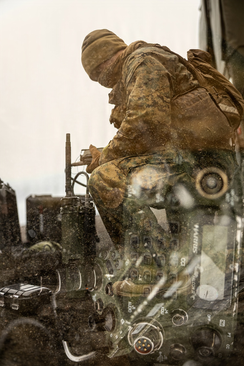 A military service member in MARPAT (Marine digital camouflage) uniform and brown watch cap is positioned on or near a military vehicle, appearing to operate or inspect equipment. The image is shot through a rain-spattered or dirty transparent surface, creating a layered reflection effect that reveals radio/communications equipment in the foreground. The visible equipment includes what appears to be a Harris Falcon III military radio system.