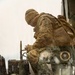 A military service member in MARPAT (Marine digital camouflage) uniform and brown watch cap is positioned on or near a military vehicle, appearing to operate or inspect equipment. The image is shot through a rain-spattered or dirty transparent surface, creating a layered reflection effect that reveals radio/communications equipment in the foreground. The visible equipment includes what appears to be a Harris Falcon III military radio system.