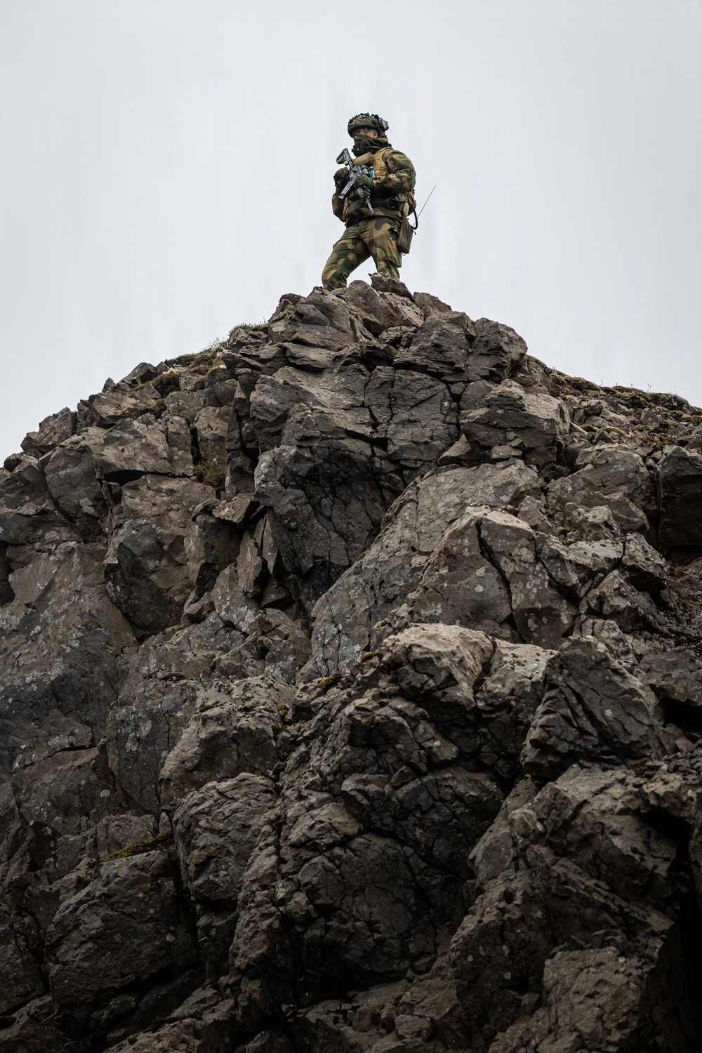 A single armed soldier in camouflage uniform stands atop a rugged, jagged rocky outcrop against an overcast sky. The soldier is equipped with full tactical gear including a helmet, body armor, rifle, and radio antenna. The low-angle perspective emphasizes the height and dominance of the position.