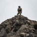 A single armed soldier in camouflage uniform stands atop a rugged, jagged rocky outcrop against an overcast sky. The soldier is equipped with full tactical gear including a helmet, body armor, rifle, and radio antenna. The low-angle perspective emphasizes the height and dominance of the position.