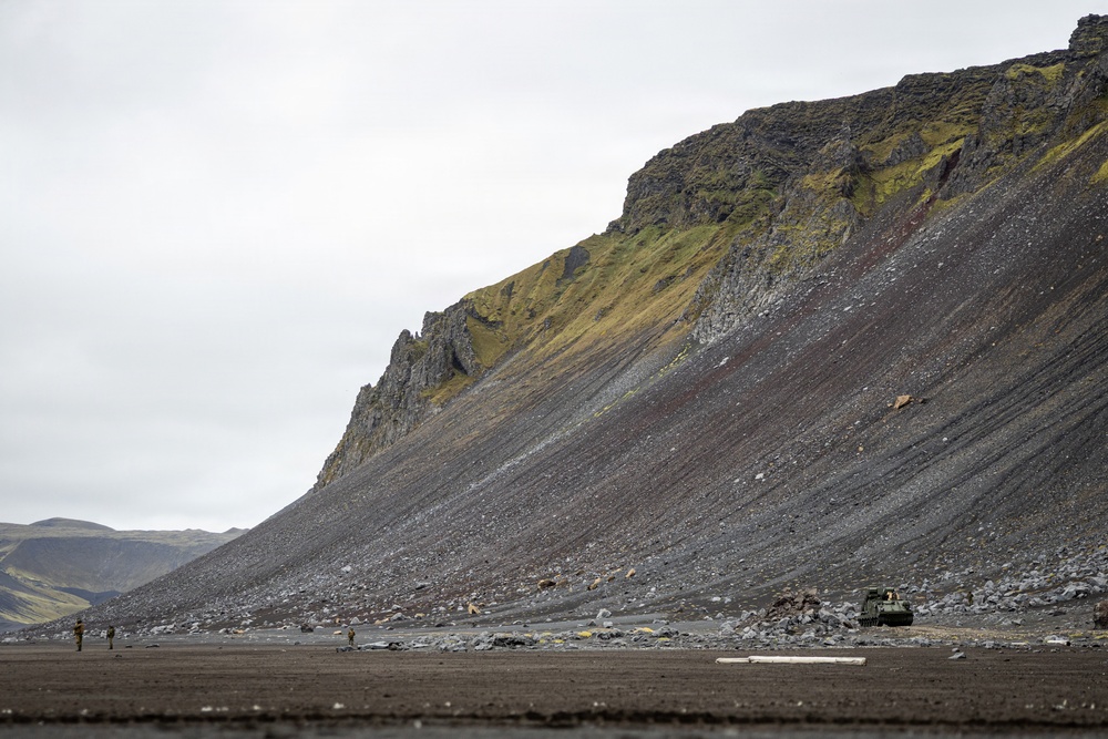 A dramatic volcanic landscape featuring a large dark scree-covered mountain slope with green moss patches near the summit. At the base of the slope, several small human figures and what appears to be a military or heavy-duty vehicle are visible, providing scale to the massive terrain. The overcast sky and dark basaltic rock suggest an Icelandic or similar sub-arctic volcanic environment.