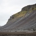 A dramatic volcanic landscape featuring a large dark scree-covered mountain slope with green moss patches near the summit. At the base of the slope, several small human figures and what appears to be a military or heavy-duty vehicle are visible, providing scale to the massive terrain. The overcast sky and dark basaltic rock suggest an Icelandic or similar sub-arctic volcanic environment.