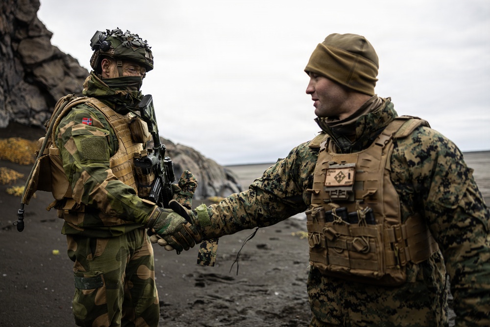 Two military personnel in full combat gear are shaking hands on a dark volcanic or rocky terrain under an overcast sky. The soldier on the left wears a Norwegian flag patch and a camouflage helmet with face covering, while the soldier on the right wears MARPAT (US Marine Corps) digital camouflage with a tan beanie. The setting and attire suggest a joint military exercise or allied forces meeting.