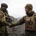 Two military personnel in full combat gear are shaking hands on a dark volcanic or rocky terrain under an overcast sky. The soldier on the left wears a Norwegian flag patch and a camouflage helmet with face covering, while the soldier on the right wears MARPAT (US Marine Corps) digital camouflage with a tan beanie. The setting and attire suggest a joint military exercise or allied forces meeting.
