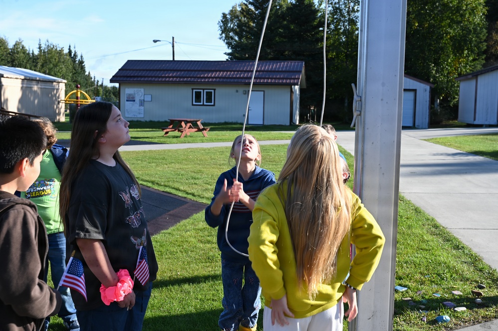 A group of children are gathered around a flagpole outdoors, appearing to raise or lower a flag. One child in a blue hoodie is pulling the rope on the flagpole while others watch, and at least one child is holding small American flags. The setting appears to be a school or community facility with buildings, a playground, and well-maintained grass visible in the background.