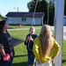 A group of children are gathered around a flagpole outdoors, appearing to raise or lower a flag. One child in a blue hoodie is pulling the rope on the flagpole while others watch, and at least one child is holding small American flags. The setting appears to be a school or community facility with buildings, a playground, and well-maintained grass visible in the background.