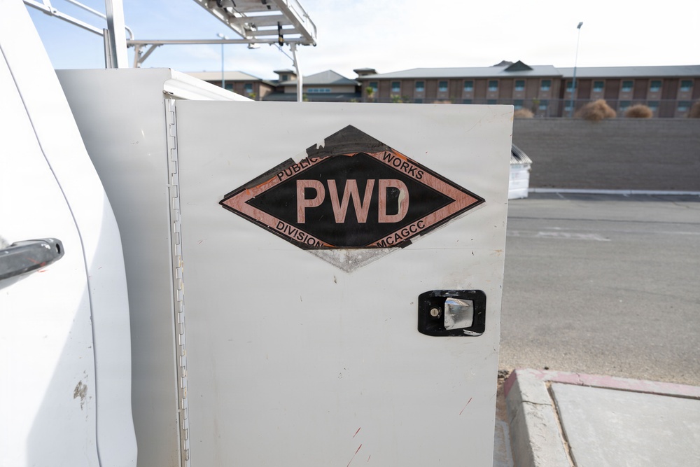 The image shows a close-up of a white utility truck or service vehicle storage compartment door bearing a 'PWD' (Public Works Division) diamond-shaped logo. The logo includes the text 'PUBLIC WORKS' at the top and 'DIVISION MCAGCC' at the bottom, indicating this vehicle belongs to the Marine Corps Air Ground Combat Center (MCAGCC). The vehicle is parked in what appears to be a paved lot near a multi-story brick building.