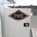The image shows a close-up of a white utility truck or service vehicle storage compartment door bearing a 'PWD' (Public Works Division) diamond-shaped logo. The logo includes the text 'PUBLIC WORKS' at the top and 'DIVISION MCAGCC' at the bottom, indicating this vehicle belongs to the Marine Corps Air Ground Combat Center (MCAGCC). The vehicle is parked in what appears to be a paved lot near a multi-story brick building.