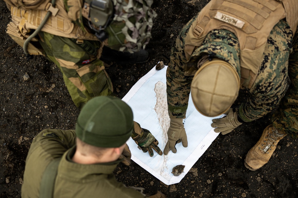 An overhead view of three military personnel in different camouflage uniforms gathered around a topographic map laid on dark ground. One soldier wearing MARPAT (US Marine Corps) digital camouflage with a name tape reading 'MURDEK' is pointing at the map alongside two others in what appears to be Swedish or Norwegian woodland camouflage. The group appears to be conducting joint military map reading or mission planning.