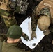 An overhead view of three military personnel in different camouflage uniforms gathered around a topographic map laid on dark ground. One soldier wearing MARPAT (US Marine Corps) digital camouflage with a name tape reading 'MURDEK' is pointing at the map alongside two others in what appears to be Swedish or Norwegian woodland camouflage. The group appears to be conducting joint military map reading or mission planning.