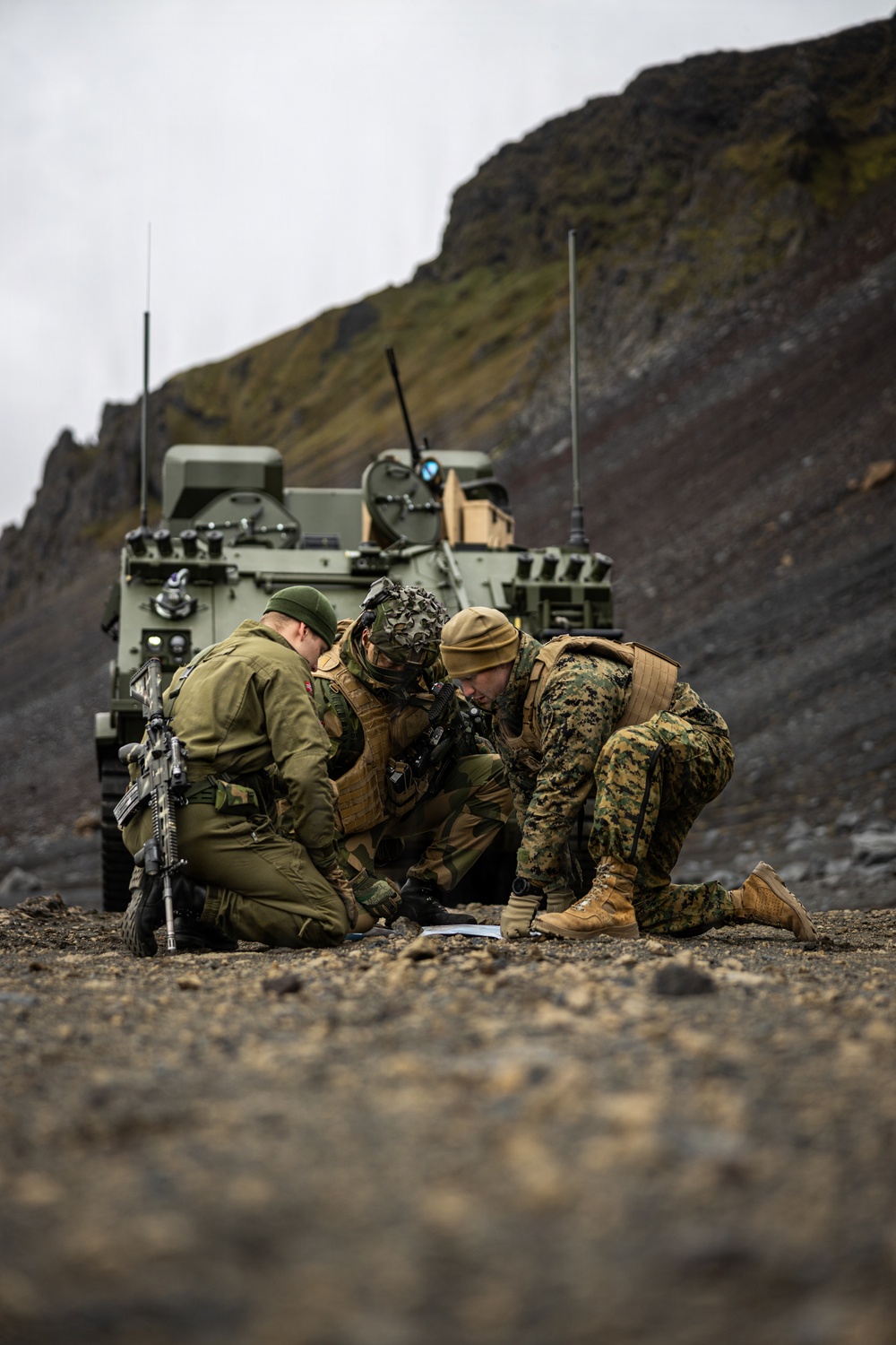 Three military personnel in mixed uniforms are crouched on rocky ground examining a document or map, with two armored military vehicles positioned behind them against a steep, rocky hillside. The soldiers appear to represent at least two different nations based on their distinct uniform patterns and equipment. The setting suggests a field exercise or operational planning scenario in a rugged, cold-weather environment.