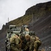 Three military personnel in mixed uniforms are crouched on rocky ground examining a document or map, with two armored military vehicles positioned behind them against a steep, rocky hillside. The soldiers appear to represent at least two different nations based on their distinct uniform patterns and equipment. The setting suggests a field exercise or operational planning scenario in a rugged, cold-weather environment.