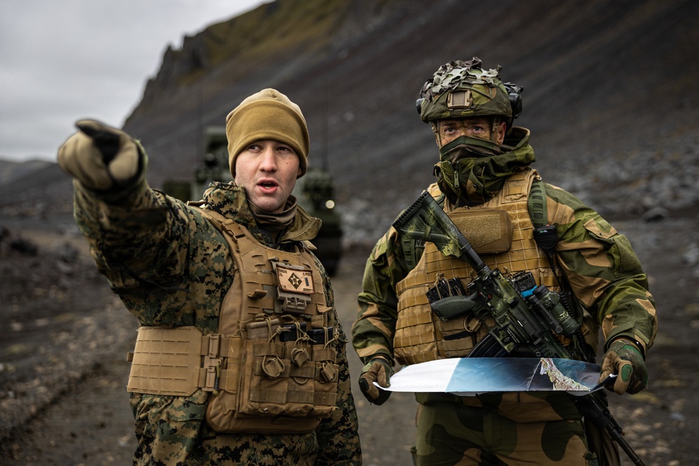 Two military personnel in full combat gear are conducting what appears to be a field coordination exercise in a rocky, mountainous terrain under overcast skies. The soldier on the left, wearing MARPAT digital camouflage consistent with US Marine Corps uniform, is pointing in a direction while the soldier on the right, wearing woodland camouflage consistent with Nordic/Norwegian military pattern, holds a map and carries a rifle. A military vehicle is visible in the background.
