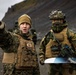 Two military personnel in full combat gear are conducting what appears to be a field coordination exercise in a rocky, mountainous terrain under overcast skies. The soldier on the left, wearing MARPAT digital camouflage consistent with US Marine Corps uniform, is pointing in a direction while the soldier on the right, wearing woodland camouflage consistent with Nordic/Norwegian military pattern, holds a map and carries a rifle. A military vehicle is visible in the background.