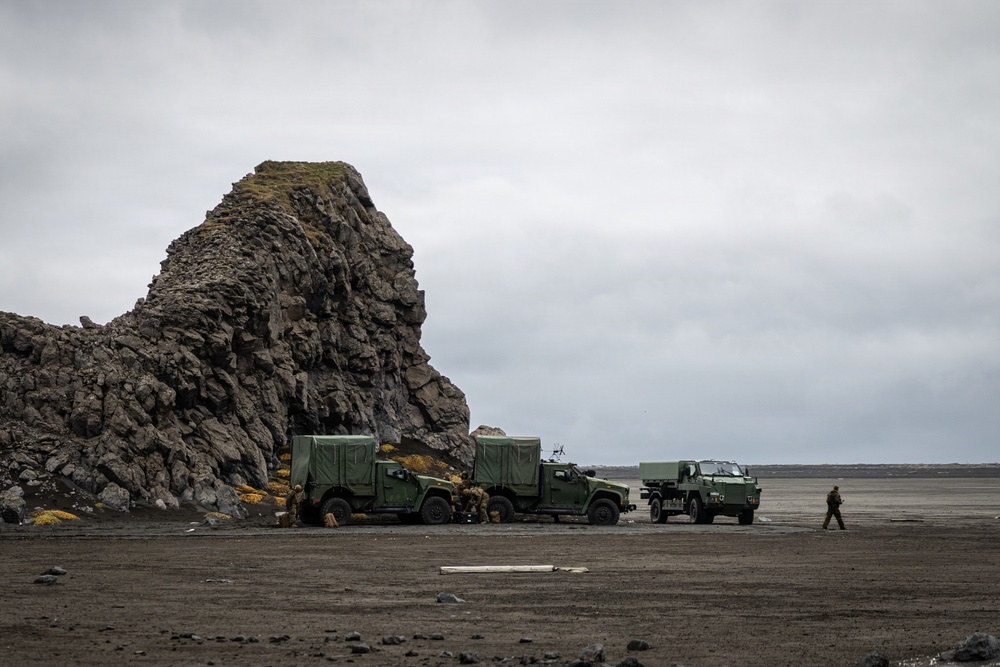 Three military tactical vehicles in olive/dark green livery are positioned on a dark volcanic beach or coastal plain adjacent to a large basalt rock formation. Several personnel in military uniforms are visible around the vehicles, appearing to conduct maintenance or operational activities. The overcast sky and dark sandy terrain suggest a northern or subarctic coastal environment.