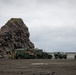 Three military tactical vehicles in olive/dark green livery are positioned on a dark volcanic beach or coastal plain adjacent to a large basalt rock formation. Several personnel in military uniforms are visible around the vehicles, appearing to conduct maintenance or operational activities. The overcast sky and dark sandy terrain suggest a northern or subarctic coastal environment.