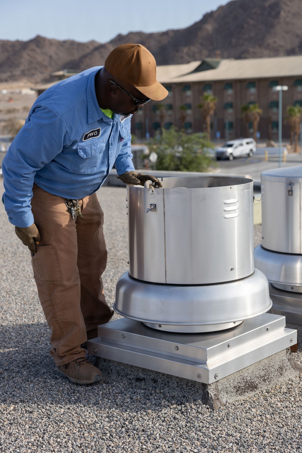 A maintenance worker wearing a blue 'PWD' uniform shirt and tan cap is inspecting or servicing a rooftop exhaust fan/ventilation unit on a flat gravel roof. The setting appears to be a desert environment, with arid mountains and a multi-story building visible in the background. A second similar ventilation unit is partially visible to the right.