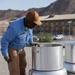 A maintenance worker wearing a blue 'PWD' uniform shirt and tan cap is inspecting or servicing a rooftop exhaust fan/ventilation unit on a flat gravel roof. The setting appears to be a desert environment, with arid mountains and a multi-story building visible in the background. A second similar ventilation unit is partially visible to the right.