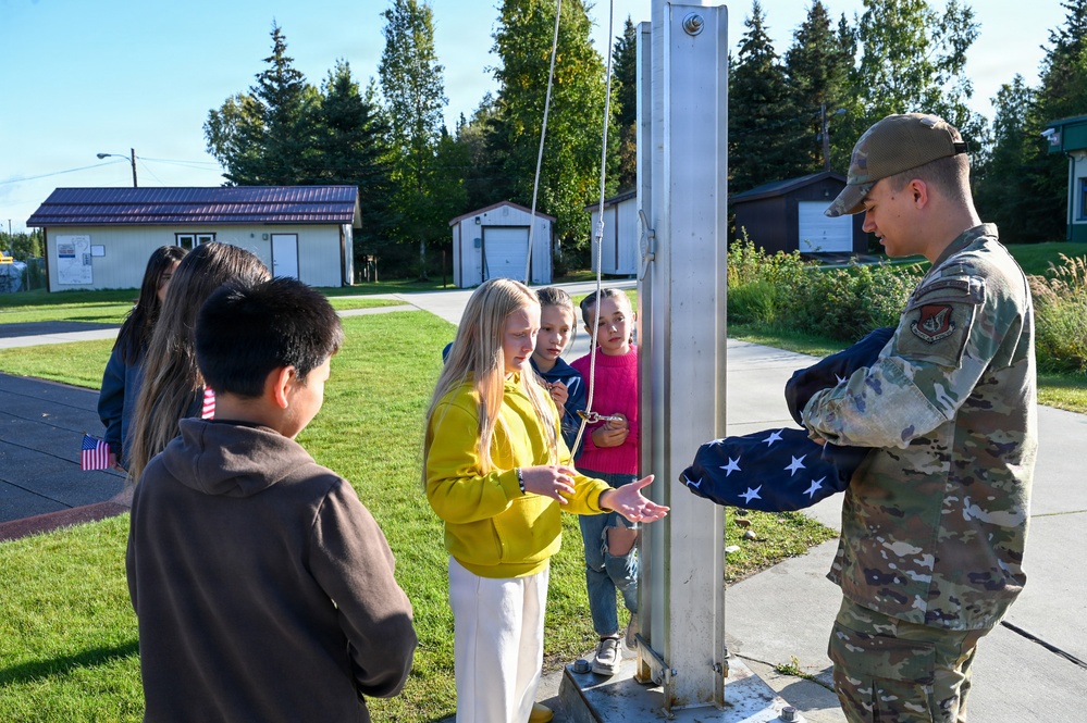 A U.S. Air Force service member in OCP (Operational Camouflage Pattern) uniform is instructing a group of children near a flagpole, holding a folded American flag. The setting appears to be a military base or school grounds on a clear, sunny day. The interaction suggests an educational demonstration about flag ceremony protocols.