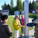 A U.S. Air Force service member in OCP (Operational Camouflage Pattern) uniform is instructing a group of children near a flagpole, holding a folded American flag. The setting appears to be a military base or school grounds on a clear, sunny day. The interaction suggests an educational demonstration about flag ceremony protocols.
