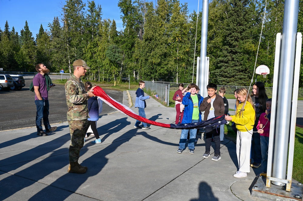 A military service member in camouflage uniform is teaching a group of school-age children how to properly handle and fold an American flag near a flagpole. The scene takes place on a school or base playground on a sunny day, with multiple flagpoles visible and a basketball hoop in the background. Two adult civilians observe the activity from the left side.