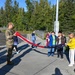 A military service member in camouflage uniform is teaching a group of school-age children how to properly handle and fold an American flag near a flagpole. The scene takes place on a school or base playground on a sunny day, with multiple flagpoles visible and a basketball hoop in the background. Two adult civilians observe the activity from the left side.
