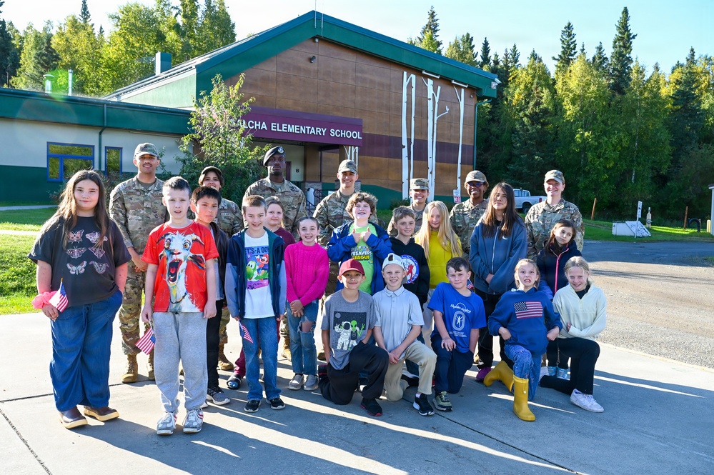 A group of elementary school children pose with several military personnel in camouflage uniforms in front of a building identified as '[A]LCHA ELEMENTARY SCHOOL.' The photo appears to be taken during a sunny day, likely during a military community outreach or school visit event. Some children are holding small American flags, suggesting a patriotic or ceremonial occasion.