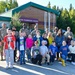 A group of elementary school children pose with several military personnel in camouflage uniforms in front of a building identified as '[A]LCHA ELEMENTARY SCHOOL.' The photo appears to be taken during a sunny day, likely during a military community outreach or school visit event. Some children are holding small American flags, suggesting a patriotic or ceremonial occasion.