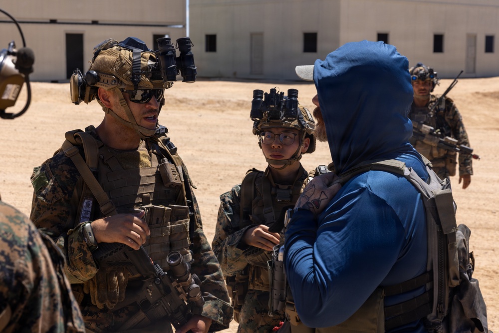 The image shows a group of military personnel in full combat gear, including helmets with night vision device mounts, tactical vests, and camouflage uniforms, engaged in a conversation with a civilian or role-player wearing a blue hoodie and white cap. The setting appears to be a military training facility or mock urban environment with sand-covered ground and white building structures in the background. This appears to be a training exercise or briefing scenario.