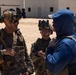 The image shows a group of military personnel in full combat gear, including helmets with night vision device mounts, tactical vests, and camouflage uniforms, engaged in a conversation with a civilian or role-player wearing a blue hoodie and white cap. The setting appears to be a military training facility or mock urban environment with sand-covered ground and white building structures in the background. This appears to be a training exercise or briefing scenario.