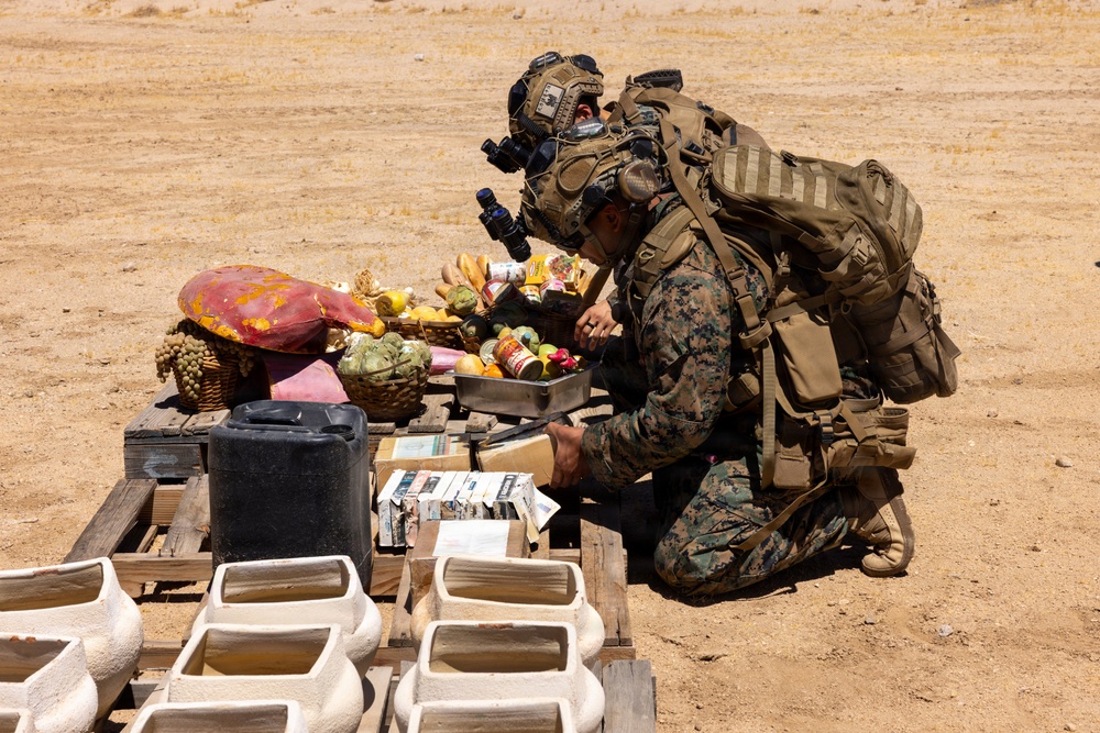 A fully equipped military personnel in MARPAT (Marine pattern) camouflage uniform is kneeling and inspecting items on a wooden pallet in an arid, desert-like environment. The scene appears to depict a search or inspection operation, with various goods including fruits, canned items, documents, and containers laid out on the pallet. White rectangular containers are arranged in rows in the foreground.