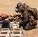 A fully equipped military personnel in MARPAT (Marine pattern) camouflage uniform is kneeling and inspecting items on a wooden pallet in an arid, desert-like environment. The scene appears to depict a search or inspection operation, with various goods including fruits, canned items, documents, and containers laid out on the pallet. White rectangular containers are arranged in rows in the foreground.