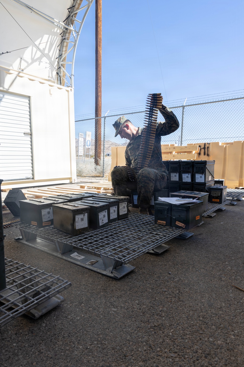 A military service member in camouflage uniform and patrol cap is seated outdoors handling a belt of linked ammunition. The individual is surrounded by numerous military ammunition cans (ammo cans) arranged on metal grating pallets. The setting appears to be a military installation or range facility with chain-link fencing and a storage structure visible in the background.
