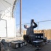 A military service member in camouflage uniform and patrol cap is seated outdoors handling a belt of linked ammunition. The individual is surrounded by numerous military ammunition cans (ammo cans) arranged on metal grating pallets. The setting appears to be a military installation or range facility with chain-link fencing and a storage structure visible in the background.