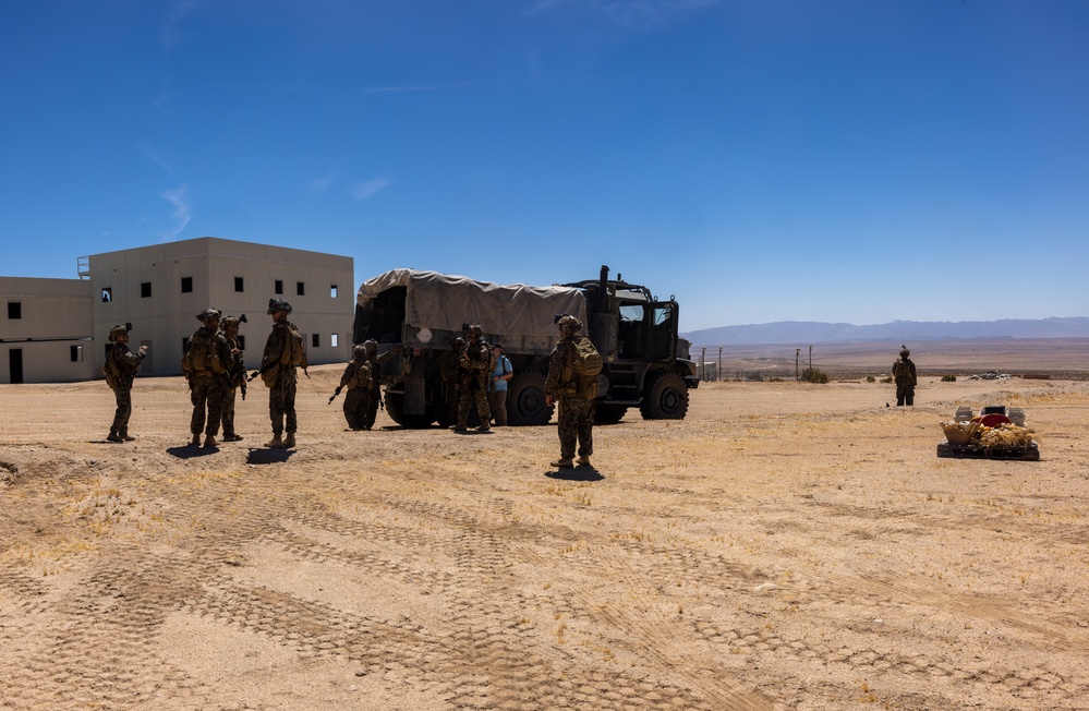 A group of military personnel in combat gear are gathered around a large canvas-covered military truck in an arid desert training environment. The scene appears to be a tactical exercise or training operation at a military installation, featuring mock urban structures in the background. The setting is consistent with a desert training facility, likely in the American Southwest based on terrain and architecture.